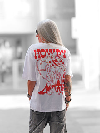 Nahaufnahme des OFF#DLY Tee Howdy white/red mit Western-Print und Glitzerdetails auf der Rückseite, lässigem Schnitt und Oversize-Charakter, präsentiert von einer Person im Freien.
