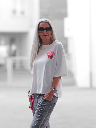 Nahaufnahme des OFF#DLY Tee Cherry offwhite T-Shirts mit auffälligem Kirsch-Design und abnehmbarem Schlüsselanhänger, getragen von einer Frau in weißem Shirt, passend für einen lässigen Sommerlook.