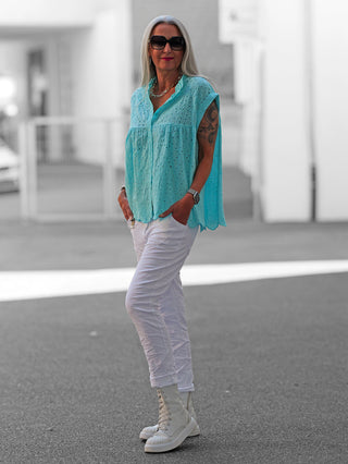 Die OFF#DLY Bluse Orea türkis zeigt eine fein bestickte Textur mit Knopfleiste und geschwungener Saumkante, getragen auf der Straße, kombiniert mit weißen Boots, betont Leichtigkeit und mediterranes Flair.