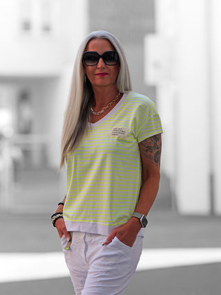 Eine Frau trägt das OFF#DLY V-Neck Ringelshirt in Weiß/Gelb mit feinen Streifen und einem charmanten Patch auf der Brust, das einen lässigen, sommerlichen Stil ausstrahlt.
