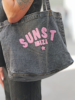 Nahaufnahme der OFF#DLY Bag Sunset Ibiza black denim, mit pinken Chenille-Buchstaben SUNSET IBIZA und kleinem Stern. Der Träger zeigt tätowierten Unterarm und schwarze Kleidung.