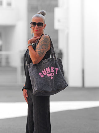 Frau mit hochgestecktem silbernem Haar trägt die geräumige OFF#DLY Bag Sunset Ibiza black denim über der Schulter, mit auffälligem pinkem Schriftzug, in urbaner Umgebung.