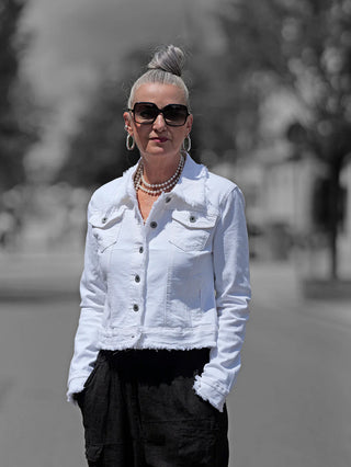 Die OFF#DLY Jacke Bologna panacotta zeigt lässigen Charme: weiße Denim-Jacke mit Fransendetails, getragen von einer Frau mit silbernem Haar, kombiniert mit schwarzen Hosen und Perlenketten.