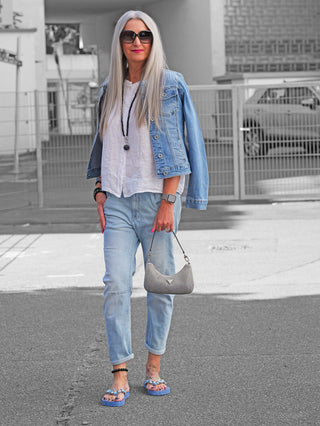 Frau trägt OFF#DLY Jacke Palermo midblue: Jeansjacke mit Strassdetails, über Schultern drapiert, kombiniert mit weißer Bluse und Jeans. Accessoires: Sonnenbrille, silberne Handtasche, Smartwatch.