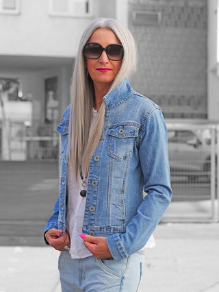 Frau in urbaner Umgebung trägt die OFF#DLY Jacke Palermo midblue, eine Jeansjacke mit Strassdetails. Kombiniert mit weißem T-Shirt und passender Jeans, betont durch schwarze Sonnenbrille und pinkem Lippenstift.