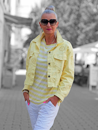 Frau trägt die OFF#DLY Jacke Lipari gelb, eine eyelet-bestickte, leicht oversize, gelbe Denim-Jacke mit aufgestelltem Kragen, über einem gestreiften Top, kombiniert mit weißen Hosen und auffälligen Accessoires.