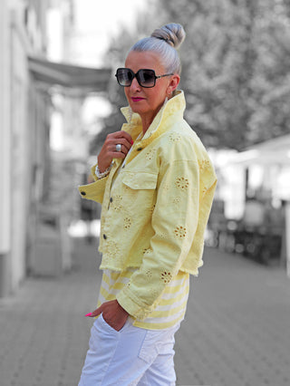 Eine stilvolle Frau in Profilansicht trägt die OFF#DLY Jacke Lipari gelb mit Lochstickerei und Pop-Kragen über einem gestreiften Oberteil, kombiniert mit weißen Hosen und auffälligen Accessoires.