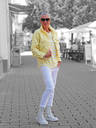 Person trägt die OFF#DLY Jacke Lipari gelb mit Lochstickerei, kombiniert mit gestreiftem T-Shirt, weißen Hosen und weißen Stiefeln. Fokus auf lässigem, sommerlichem Look der Jacke.