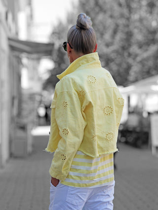 Person von hinten in einer OFF#DLY Jacke Lipari gelb mit Lochstickerei, trägt gestreiftes Shirt und weiße Hose; Haare im Dutt, Sonnenbrille. Hintergrund: verschwommene Stadtansicht.