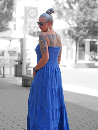 Nahaufnahme einer Frau in einem blauen OFF#DLY Kleid Bergamo mit smock-elastischem Brustbereich und stufigem Rock, kombiniert mit einer Sonnenbrille, im Freien getragen.