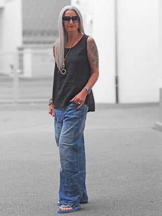 Ältere Person in OFF#DLY Wide Denim midblue, locker sitzende Jeans mit weitem Bein, Tattoos sichtbar. Kombiniert mit schwarzem ärmellosen Top und Sandalen, urbaner Hintergrund. Perfekt für lässige Looks.