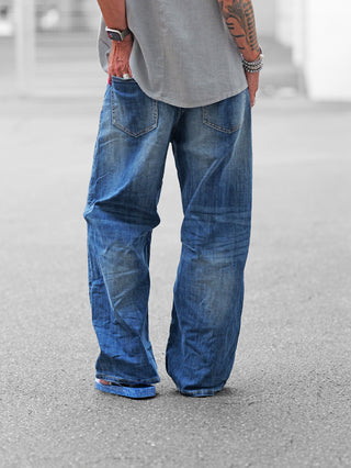 Person von hinten in OFF#DLY Wide Denim midblue, weit geschnittene blaue Jeans mit Vintage-Look. Ergänzt durch blaue Flip-Flops, graues Hemd, Smartwatch und bunte Nägel.