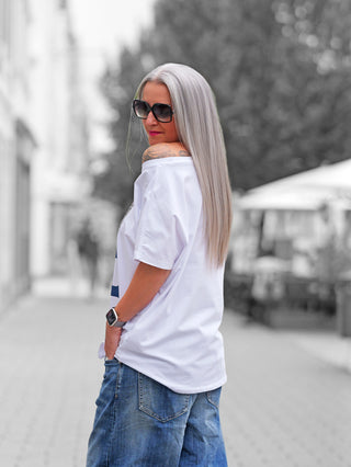 Person trägt das OFF#DLY Tee Wild white, locker über eine Schulter, mit sichtbarem Tattoo am Oberarm. Kombiniert mit großen Sonnenbrillen, Jeans und einer Smartwatch, verkörpert es lässigen urbanen Stil.