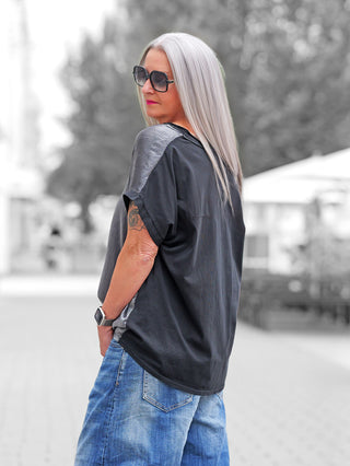 Person trägt das OFF#DLY T-Shirt Luino black mit glänzenden Schulterpanelen und goldener Aufschrift. Kombiniert mit blauen Jeans, große Sonnenbrille. Sichtbares Tattoo am Arm, stilvoll und modern.