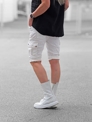 Person trägt OFF#DLY Short Lecce offwhite, kniehohe Cargo-Shorts mit seitlichen Klappentaschen und umgeschlagenen Abschlüssen, kombiniert mit hohen weißen Schnürstiefeln und schwarzem ärmellosen Oberteil auf gepflastertem Boden.