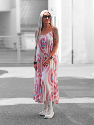 Frau trägt OFF#DLY Kleid Milano rosa/multi, ein fließendes, knöchellanges Sommerkleid mit großen, abstrakten pink-blauen Kreisen, kombiniert mit weißen Stiefeln, Sonnenbrille und perlenschmuck.