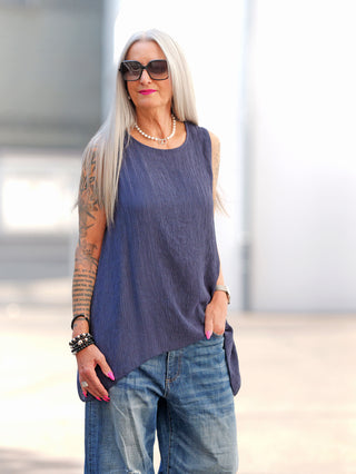 Silberhaarige Frau in OFF#DLY Top Taranto blue, ärmellos mit Crinkle-Textur, asymmetrisch drapiert, kombiniert mit lockeren Jeans, Tattoos und Schmuck.