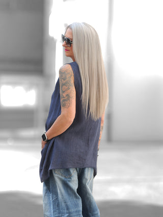 Person trägt OFF#DLY Top Taranto blue mit Crinkle-Effekt, ärmellos, verlängerte Rückseite, über light-washed Jeans. Oversized Sonnenbrille, silberblonde Haare, Tattoos am Arm, Hände in den Taschen.