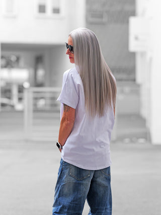 Erwachsene Person trägt das OFF#DLY Tee sneaker white mit auffälligem Sneaker-Print und goldenen Akzenten, kombiniert mit blauer Jeans. Sichtbar sind eine Smartwatch und ein Unterarmtattoo.