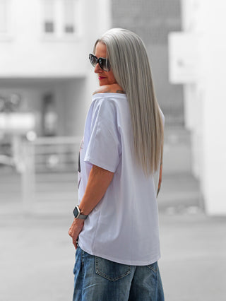 Frau mit langen, silbernen Haaren und Sonnenbrille trägt das OFF#DLY Tee Romantic white in urbaner Kulisse, locker über der Schulter fallend, kombiniert mit blauen Jeans und Smartwatch.