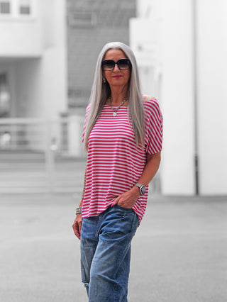 Person in urbanem Umfeld trägt das OFF#DLY Margot Stripe Tee pink, ein locker geschnittenes rosa-weiß gestreiftes Kurzarmshirt mit Rundhals und geschnittenen Kanten, kombiniert mit blauer Jeans und Accessoires.