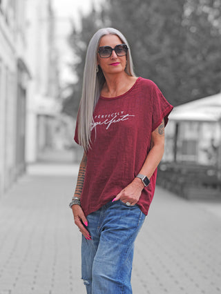 Erwachsene Frau trägt ein OFF#DLY T-Shirt Baveno bordeaux mit „Perfectly imperfect“ Schriftzug, kombiniert mit blauen Jeans und einer Smartwatch, während sie auf einer gepflasterten Straße steht.