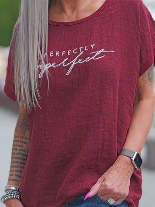 Person mit silbernem Haar trägt ein OFF#DLY T-Shirt Baveno bordeaux mit glitzerndem perfectly imperfect Schriftzug, kombiniert mit tätowierten Unterarmen, pinken Nägeln, Smartwatch und Jeans.