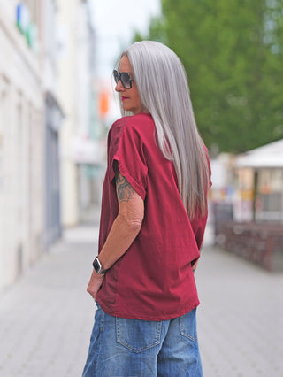 Person mit langen, silbernen Haaren trägt das OFF#DLY T-Shirt Baveno bordeaux. Locker geschnitten mit kurzen Ärmeln, „Perfectly imperfect“-Schriftzug vorne, kombiniert mit blauen Jeans und einer Smartwatch am Handgelenk.