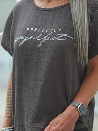 Torso mit OFF#DLY T-Shirt Baveno braun, glitzernder „Perfectly Imperfect“-Schriftzug. Sichtbar: Tattoos, Armbänder, silberne Smartwatch, lange aschblonde Haare über einer Schulter.