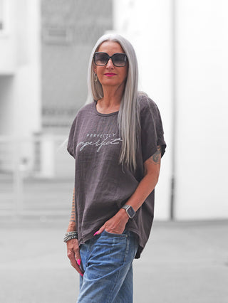 Frau trägt das OFF#DLY T-Shirt Baveno braun mit „Perfectly Imperfect“-Glitzerschrift, kombiniert mit blauen Jeans. Sie hat graues Haar, Sonnenbrille, Armbänder und eine Smartwatch.
