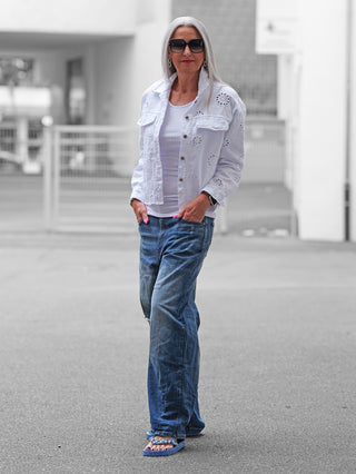 Ältere Frau trägt OFF#DLY Jacke Lipari white: weiße, augestickte Denim-Jacke über weißem Top, kombiniert mit blauen Jeans und verzierten Flip-Flops, symbolisiert entspannten Sommerstil.