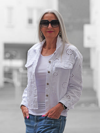 Ältere Frau in urbaner Umgebung trägt die OFF#DLY Jacke Lipari white: weiße, Lochstickerei-Denim-Jacke über einem gerippten Tanktop, kombiniert mit blauer Jeans und silbernen Ohrringen.