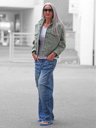Frau mit langen, silbernen Haaren trägt die OFF#DLY Jacke Modica matcha, eine olivgrüne, kurz geschnittene Jacke mit Brusttaschen, über einem weißen Tanktop kombiniert mit blauen Jeans und Flip-Flops.