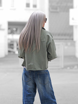 Person von hinten in urbaner Umgebung trägt die OFF#DLY Jacke Modica matcha, eine olivgrüne, kastig geschnittene Jacke mit offenen Saumkanten, kombiniert mit entspannten blauen Jeans.