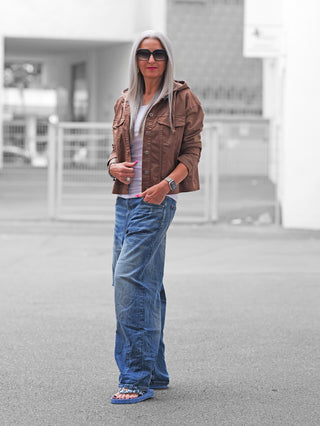 Ältere Frau in lockerem Look mit der OFF#DLY Jacke Pisogne schokobraun, die über einem weißen T-Shirt getragen wird, kombiniert mit weiten blauen Jeans und blauen Flip-Flops.