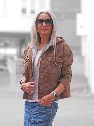Frau mit grauen Haaren trägt die OFF#DLY Jacke Pisogne schokobraun, eine verkürzte, braune Jacke mit Knöpfen und Snap-Taschen, über einem weißen Tanktop, kombiniert mit blauer Jeans und modernen Accessoires.