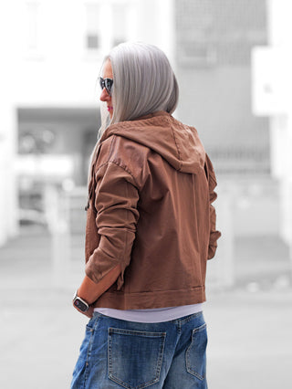 Person von hinten in urbaner Umgebung trägt die OFF#DLY Jacke Pisogne schokobraun. Die Jacke kombiniert Jeansjacken-Look mit Sweat-Elementen, Kapuze und Fransen, ideal für lässige, feminine Styles.