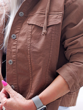 Nahaufnahme der OFF#DLY Jacke Pisogne schokobraun, getragen mit einer Hand mit pinken Nägeln. Die Jacke zeigt eine Brusttasche, Knopfleiste, Kapuzenkordel und eine silberne Smartwatch am Handgelenk.