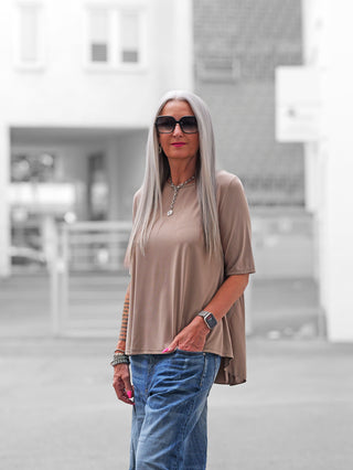 Frau trägt das OFF#DLY T-Shirt Agropoli beige mit fließendem Schnitt und einzigartiger Rückenfalte, kombiniert mit blauer Jeans, Smartwatch und Schmuck, in urbaner Umgebung.