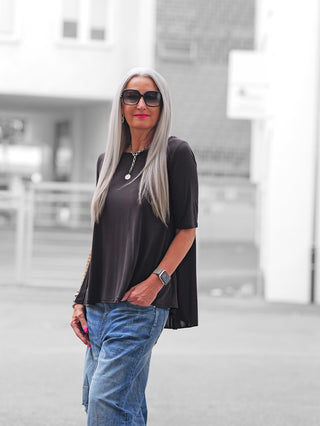 Erwachsene Frau trägt OFF#DLY T-Shirt Agropoli schwarz, locker geschnitten, mit raffiniertem Rückenteil. Kombiniert mit blauer Jeans und silberner Kette, posiert sie lässig in urbaner Umgebung.