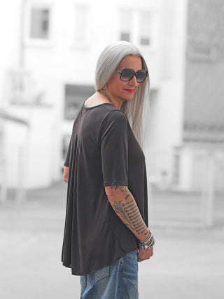 Ältere Frau in der Stadt trägt ein OFF#DLY T-Shirt Agropoli in Schwarz mit fließendem, femininen Schnitt, der am Rücken raffiniert fällt, kombiniert mit blauen Jeans und auffälligem Tattoo.