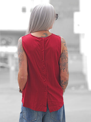 Person von hinten in urbaner Umgebung, trägt das OFF#DLY Top Follonica red. Ärmelloses, rotes Top mit Knopfleiste hinten, kombiniert mit Jeans, Tattoo-Designs auf den Armen sichtbar.