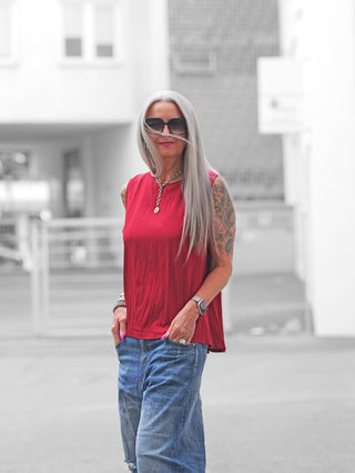 Frau mit langen silbernen Haaren trägt das ärmellose OFF#DLY Top Follonica red. Es hat eine durchgehende Knopfleiste am Rücken, kombiniert mit locker sitzenden, leicht zerrissenen blauen Jeans.