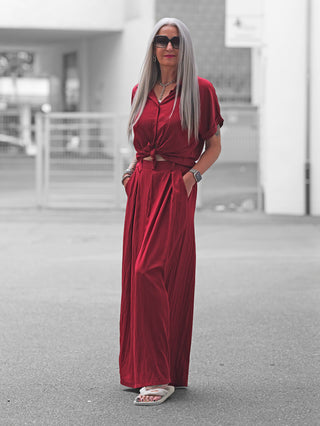 Ältere Frau trägt OFF#DLY Pant Gallipoli red, eine weite, luftige Palazzo-Hose mit elastischem Bund und seitlichen Taschen, kombiniert mit passendem rotem Oberteil und weißen Sandalen auf einer Stadtstraße.