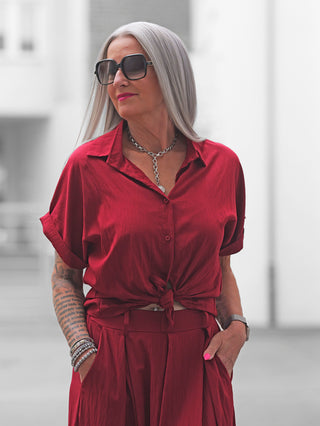 Ältere Frau trägt die OFF#DLY Bluse Soverato red, ein Statement-Stück mit leichtem Glanz, geknotet an der Taille, kombiniert mit einem silbernen Kettenarmband und Sonnenbrille.