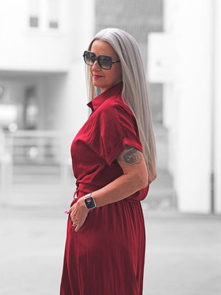 Frau in rotem Kleid OFF#DLY Bluse Soverato red mit Hemdkragen und Taillierung, trägt Sonnenbrille und Smartwatch, zeigt modernen Look mit Tattoo auf dem Arm vor urbanem Hintergrund.