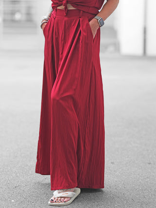 Person trägt OFF#DLY Pant Gallipoli red, eine hoch taillierte, weit geschnittene Palazzo-Hose, kombiniert mit einem passenden gebundenen Oberteil. Hände in den Taschen, ergänzt durch Armbänder und eine Smartwatch.