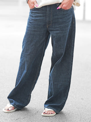 Person trägt OFF#DLY Wide Denim Bolsena dark blue, weit geschnittene Jeans mit klassischem Five-Pocket-Design, auf gepflastertem Boden. Weiße Flip-Flops und pink lackierte Fingernägel sind sichtbar.