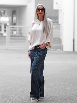 Frau trägt OFF#DLY Wide Denim Bolsena dark blue mit weitem Bein und klassischem Schnitt. Kombiniert mit lockerer Bluse, Sandalen, silbernem Schmuck und Sonnenbrille, vermittelt sie lässigen City-Style.