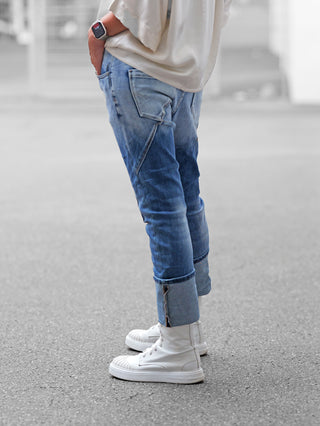 Person trägt OFF#DLY Denim Stresa midblue, locker geschnittene, umgeschlagene Jeans mit Teilungsnähten und Reißverschluss am Beinabschluss. Kombiniert mit weißen High-Top-Sneakern und lockerem Oberteil.
